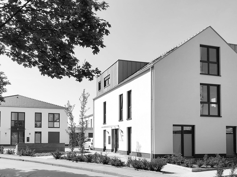 Modernes Einfamilienhaus mit einem Satteldach, großen Fenstern und einer gepflegten Gartenlandschaft, aufgenommen bei sonnigem Wetter.