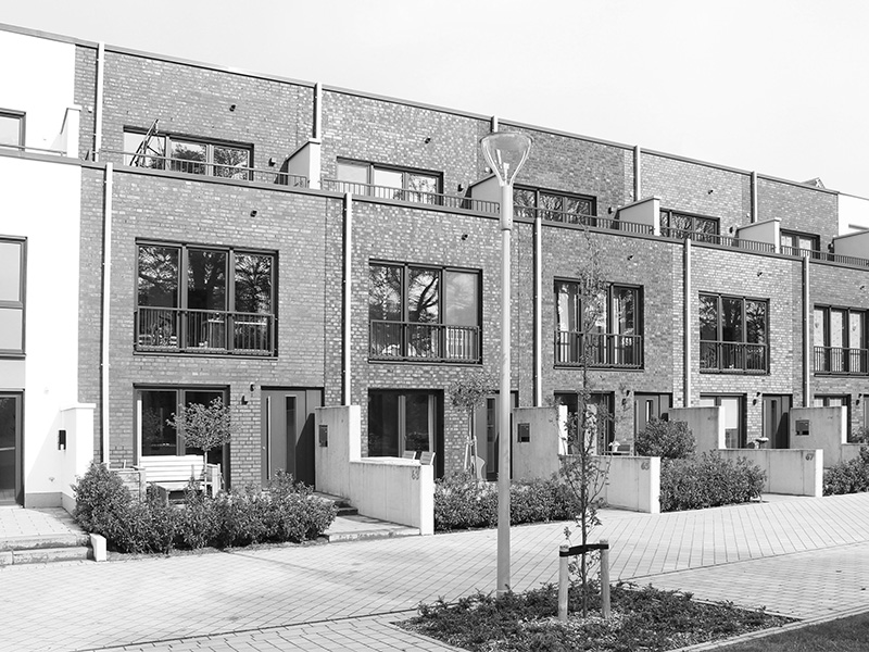 Moderne Reihenhäuser aus Backstein mit großen Fenstern und kleinen Vorgärten, die in einer ordentlichen Reihe entlang einer gepflasterten Straße stehen.